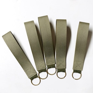 May include: Five olive green fabric keychains with gold-colored metal rings. The keychains are arranged on a white surface, with each strap extending upwards. The fabric has a textured appearance.