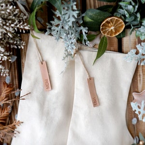 May include: Two white canvas stockings with brown leather tags. The tags are labeled "MOM" and "FRI". The stockings are surrounded by dried citrus fruit and greenery.