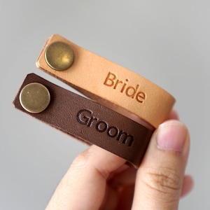 May include: Two leather bracelets with metal clasps. One is brown and says "Groom" and the other is tan and says "Bride".