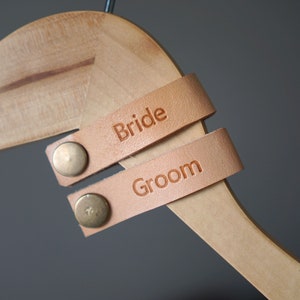 May include: Two brown leather straps with metal rivets, attached to a wooden hanger. The straps are labeled "Bride" and "Groom".