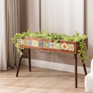 Op de afbeelding: Een rechthoekige houten plantenbak met decoratieve tegels en lange, slanke poten. De plantenbak is gevuld met groene planten en staat in een woonkamer. De tegels hebben kleurrijke geometrische patronen.