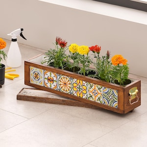 May include: A wooden planter box with colorful, patterned tiles, holding several potted flowers with orange, yellow, and red blooms. A small black pot with an orange flower, a white spray bottle, and gardening tools are nearby.