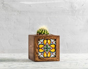 May include: A small, round cactus in a square wooden planter. The planter features a colorful, patterned tile on the front with yellow, green, and red accents. The wood has a warm, brown tone, and the background is a textured, light gray.