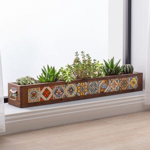 Op de afbeelding: Een rechthoekige houten plantenbak gevuld met diverse vetplanten en een kleine cactus. De bak is versierd met kleurrijke, patroon tegels langs de voorkant. Het heeft kleine metalen handvatten en staat op een witte vensterbank.