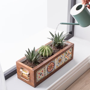 May include: A rectangular wooden planter box with a decorative tile design, filled with soil and three succulents. A person is watering the plants with a teal watering can. The planter has a brass handle.