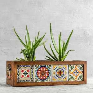Op de afbeelding: Een rechthoekige houten plantenbak met kleurrijke, patroon tegels aan de voorkant. De tegels hebben bloemen en geometrische ontwerpen in rood, blauw, geel en groen. Twee aloë vera planten met lange, groene bladeren zijn in de bak geplant.