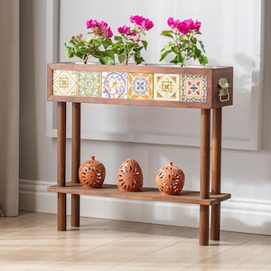 Può includere: Una consolle in legno con frontale in piastrelle decorative, che ospita piante in vaso con vibranti fiori rosa. Sotto, tre vasi in terracotta con intricati disegni traforati poggiano su un ripiano inferiore.