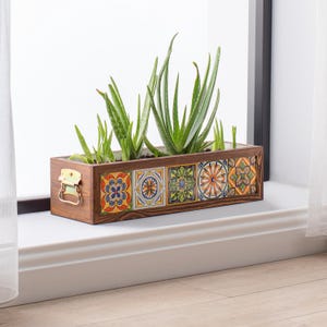 Könnte beinhalten: Ein rechteckiger Pflanzkasten aus Holz mit bunten, gemusterten Fliesen und einem Metallgriff. Die Box ist mit grünen Aloe Vera Pflanzen gefüllt. Der Pflanzkasten steht auf einer weißen Fensterbank, im Hintergrund ist ein transparenter Vorhang zu sehen.