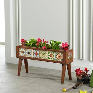 Op de afbeelding: Een houten plantenbak met kleurrijke tegelaccenten en vier schuine poten. De plantenbak is gevuld met levendig groen blad en roze en witte bloemen. Een decoratieve handgreep is zichtbaar aan de zijkant.