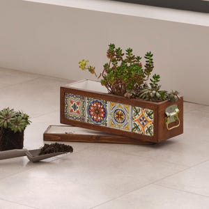 Puede incluir: Una jardinera rectangular de madera con detalles de azulejos coloridos, llena de suculentas verdes. La jardinera tiene un asa con bisagras y una base a juego. Una pequeña pala con tierra está en primer plano.