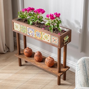 May include: A wooden planter with a rectangular design, featuring colourful patterned tiles along the front. The planter holds vibrant pink flowering plants. Below, a shelf displays three decorative, terracotta-coloured orbs.