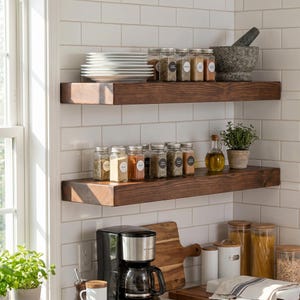 May include: Two dark brown wooden floating shelves mounted on a white subway tile wall. The top shelf holds a stack of white plates, several glass spice jars with labels, and a grey stone mortar and pestle. The bottom shelf features more spice jars.