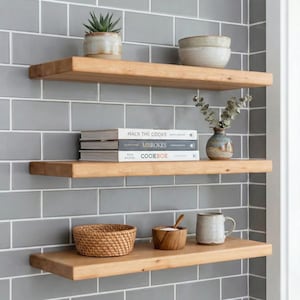 May include: Three wooden floating shelves against a gray tiled wall. The shelves hold various items, including a small potted plant, bowls, books, a vase with greenery, a woven basket, a wooden bowl, and a mug. The books have the titles "Macr the Cooks", "Abreokes", and "Cookbook".