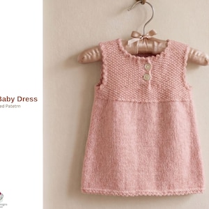 Puede incluir: Vestido de bebé tejido de color rosa claro con un corpiño texturizado y una falda lisa, colgado de una percha acolchada. El vestido tiene dos botones y un escote festoneado. El texto "Simay Baby Dress Knitted Pattern" es visible.