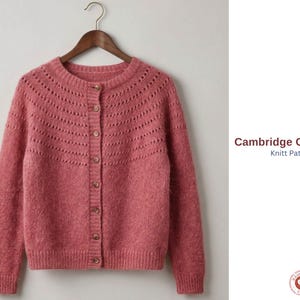 May include: A rose-pink knitted cardigan with a round neck and button closure. The Cambridge Cardigan pattern is displayed on the right. The sweater is suspended from a wooden hanger against a plain backdrop.