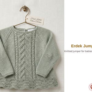 Può includere: Un maglione per neonati lavorato a maglia verde salvia con maniche lunghe, appeso a un gancio di ottone. Il maglione presenta un design centrale dettagliato e un orlo smerlato. L'etichetta recita "Hand-Knitted Artisan Collection". Il testo dice "Erdek Jumper, Knitted jumper for babies and children".