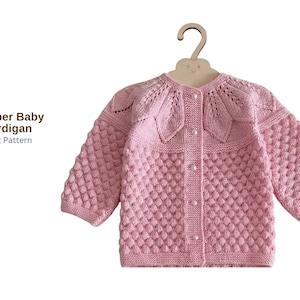 May include: A light pink baby cardigan with a textured, bubble-like knit pattern. The cardigan has a decorative floral design around the neckline and is buttoned down the front. The text "Rainber Baby Cardigan Knitt Pattern" is visible.