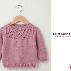 May include: A dusty rose-colored knitted baby sweater with a decorative openwork yoke, hanging on a wooden hanger. The sweater has long sleeves and a ribbed hem and cuffs. The text "Estee Spring Pullover" and "Knitted jumper for babies and children" is visible.