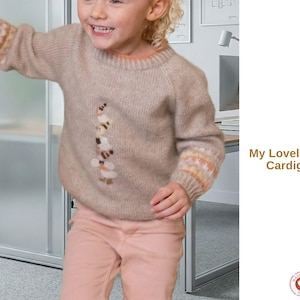 Op de afbeelding: Een beige trui met een kleurrijk motief en mouwen met patronen. De tekst "My Lovely Motif Cardigan" wordt rechts in de afbeelding weergegeven. De trui wordt door een kind gedragen.