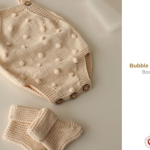May include: A cream-colored knitted bubble romper with wooden buttons and matching booties. The romper has a textured, dotted pattern. The text "Bubble Romper Booties" is visible in the image.
