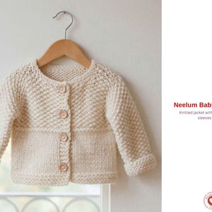 May include: Cream-coloured knitted baby jacket with seamless sleeves, hanging on a wooden hanger. The jacket has a round neckline, long sleeves, and wooden buttons. The text "Neelum Baby Jacket" is visible.