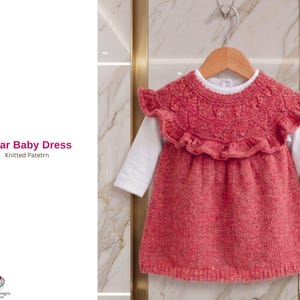 May include: A coral-colored knitted baby dress with a ruffled yoke, hanging on a wooden hanger. The dress is layered over a white long-sleeved shirt. The text "Ebrar Baby Dress Knitted Pattern" is visible on the left.