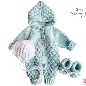 May include: A light blue knitted baby romper set with a hooded jumpsuit, bonnet, and booties. The set features a textured popcorn stitch design and wooden buttons. The text "Greenland Popcorn Romper Set" is visible.