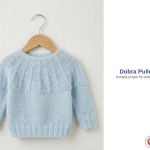 Puede incluir: Un suéter de bebé tejido de color azul claro con un diseño de canesú texturizado, colgado de una percha de madera. El suéter tiene mangas largas y un dobladillo acanalado. El texto a la derecha dice "Dobra Pullover" y "Knitted jumper for baby and kids."