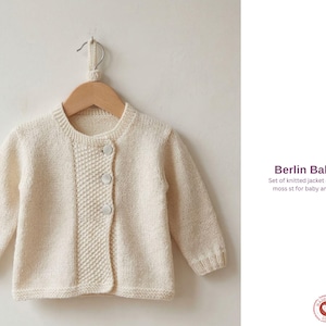 Puede incluir: Chaqueta de bebé de punto color crema con tres botones, colgada de una percha de madera. La chaqueta tiene un diseño texturizado en el panel frontal. El texto "Berlin Baby Set" es visible.