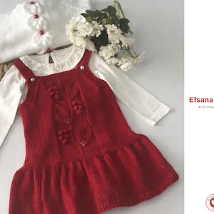 Puede incluir: Un conjunto de bebé de punto rojo que incluye un vestido con diseños de hojas y bayas, y un cárdigan blanco con flores. Debajo, una camisa blanca de manga larga. El conjunto se presenta con flores blancas.