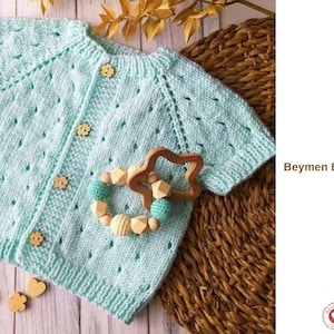 May include: A pale blue knitted baby vest with short sleeves and wooden buttons. The vest features a textured pattern and is shown with a wooden star-shaped teether and a beaded teething ring. The text "Beymen Baby Vest" is visible.