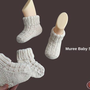 Può includere: Scarpine da neonato lavorate a maglia color crema con risvolto. Le scarpe sono esposte su sfondo marrone, con la scritta "Muree Baby Shoes" in bianco. Le scarpe sono fatte a mano e sembrano morbide e calde.