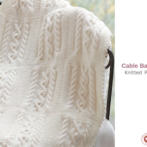 Op de afbeelding: Crèmekleurige kabelgebreide deken over een stoel gedrapeerd. De deken heeft een herhalend patroon van gevlochten kabelontwerpen en getextureerde steken. De woorden "Cable Balanket Knitted Patetrn" zijn zichtbaar.