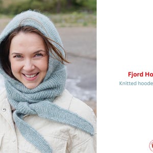 Può includere: Sciarpa con cappuccio lavorata a maglia blu chiaro, con il testo "Fjord Hood" e "Knitted hooded scarf". La sciarpa è indossata come un cappuccio e ha lunghi lacci. La persona nella foto indossa una giacca bianca.