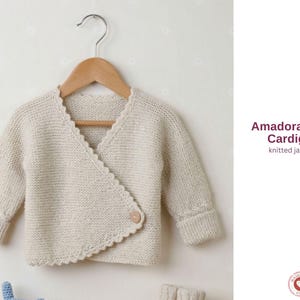 May include: A cream-colored baby cardigan with a scalloped edge and a single button closure. The knitted jacket is displayed on a wooden hanger. The text "Amadora Baby Cardigan" and "knitted jacket" are visible.