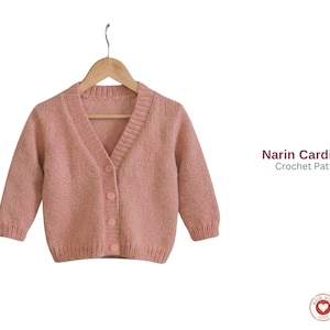 Könnte beinhalten: Ein hellrosa Cardigan mit Knöpfen, der an einem Holzbügel hängt. Der Pullover hat einen V-Ausschnitt und drei Knöpfe. Der Text "Narin Cardigan Crochet Pattern" ist auf der rechten Seite des Bildes zu sehen.