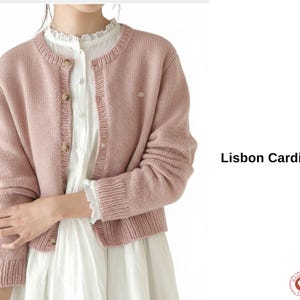 May include: A light pink Lisbon cardigan with a round neckline and button closure. The long-sleeved sweater is made of a knitted material and is shown over a white blouse. The text "Lisbon Cardigan" is displayed on the right.
