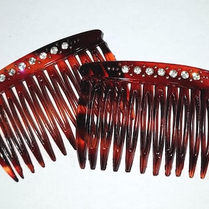 Könnte beinhalten: Zwei Haarkämme aus Schildpatt mit gewölbter Oberseite und langen Zähnen. Jeder Kamm ist mit einer Reihe funkelnder Strasssteine verziert. Die Kämme haben eine warme braune Farbe mit dunkleren braunen Marmorierungen.