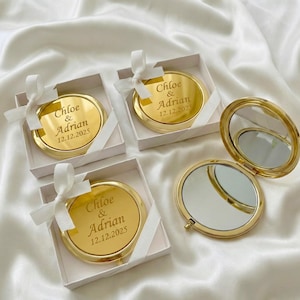 以下が含まれることがあります： 「Chloe & Adrian」と日付「12.12.2025」が刻まれたゴールドのコンパクトミラー。白い箱に入っており、白いリボンで結ばれています。1つのミラーは開いており、反射面が見えています。