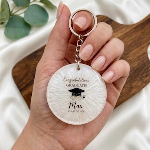 Pode incluir: Um chaveiro de formatura branco e redondo com um porta-chaves prateado. O chaveiro apresenta o texto "Parabéns Graduado!" acima de um gráfico de chapéu de formatura e o nome "Max" com "Turma de 2026" abaixo.