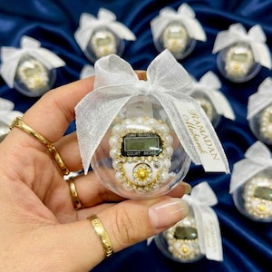 May include: Clear plastic ornament with a pearl and gold embellished digital counter. The ornament is tied with a white ribbon and a tag that reads "Ramadan Mubarak". Other similar ornaments are in the background.