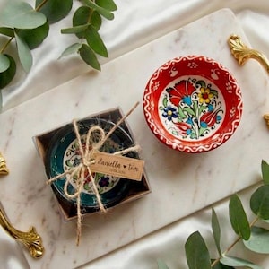 Op de afbeelding: Een rood-witte bloemmotief keramische kom en een set teal en bloemmotief kommen in een doorzichtige doos, vastgebonden met touw. De kommen staan op een witte marmeren dienblad met gouden handvatten, omringd door groen.