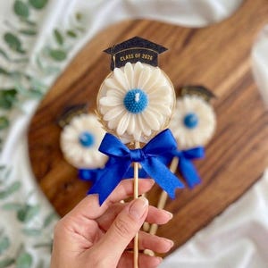May include: Graduation-themed daisy-shaped decorations. Each decoration features a white daisy with a blue center, a gold wire frame, and a blue ribbon. A small black graduation cap with "CLASS OF 2026" is attached to the top.