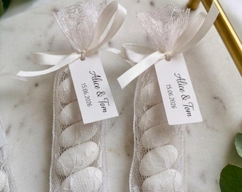May include: Two vertical white lace favor bags filled with white almond candies. Each bag is tied with a white satin ribbon and features a rectangular white paper tag printed with the text Alice & Tom and the date 15.06.2026 in black ink.