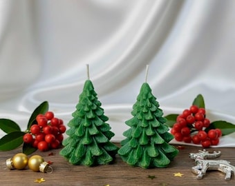 May include: Two green Christmas tree-shaped candles with white wicks. The candles are on a wooden surface, decorated with gold ornaments, red berries, and a silver reindeer figurine. The background is a white satin cloth.