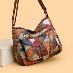 Peut inclure: Sac bandoulière patchwork multicolore avec une bandoulière marron réglable. Le sac présente des formes géométriques dans les tons orange, bleu, rouge et blanc. Il possède plusieurs compartiments zippés et un détail en cuir tressé.