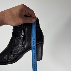 Pu&ograve; includere: Stivaletto alla caviglia in pelle nera con zip laterale e tacco a blocco. Un metro a nastro blu mostra l'altezza dello stivale. L'altezza dello stivale &egrave; di circa 20 cm.