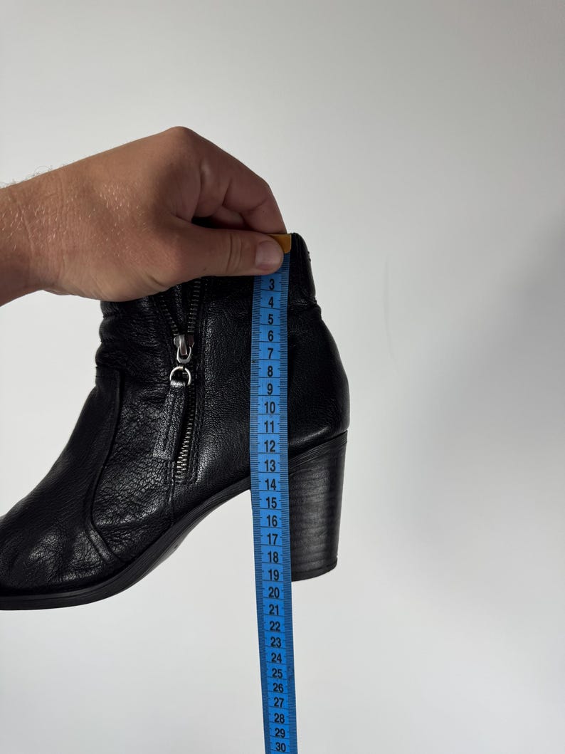 Pu&ograve; includere: Stivaletto alla caviglia in pelle nera con zip laterale e tacco a blocco. Un metro a nastro blu mostra l'altezza dello stivale. L'altezza dello stivale &egrave; di circa 20 cm.