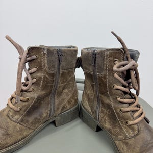 K&ouml;nnte beinhalten: Ein Paar braune Stiefeletten mit abgenutzter Lederoptik, Schn&uuml;rung mit passenden braunen Schn&uuml;rsenkeln, messingfarbenen &Ouml;sen und vertikalen seitlichen Rei&szlig;verschl&uuml;ssen. Die Stiefel haben einen niedrigen Absatz und sichtbare N&auml;hte an den Seiten.