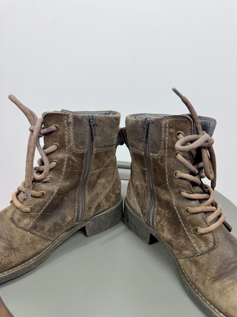 K&ouml;nnte beinhalten: Ein Paar braune Stiefeletten mit abgenutzter Lederoptik, Schn&uuml;rung mit passenden braunen Schn&uuml;rsenkeln, messingfarbenen &Ouml;sen und vertikalen seitlichen Rei&szlig;verschl&uuml;ssen. Die Stiefel haben einen niedrigen Absatz und sichtbare N&auml;hte an den Seiten.